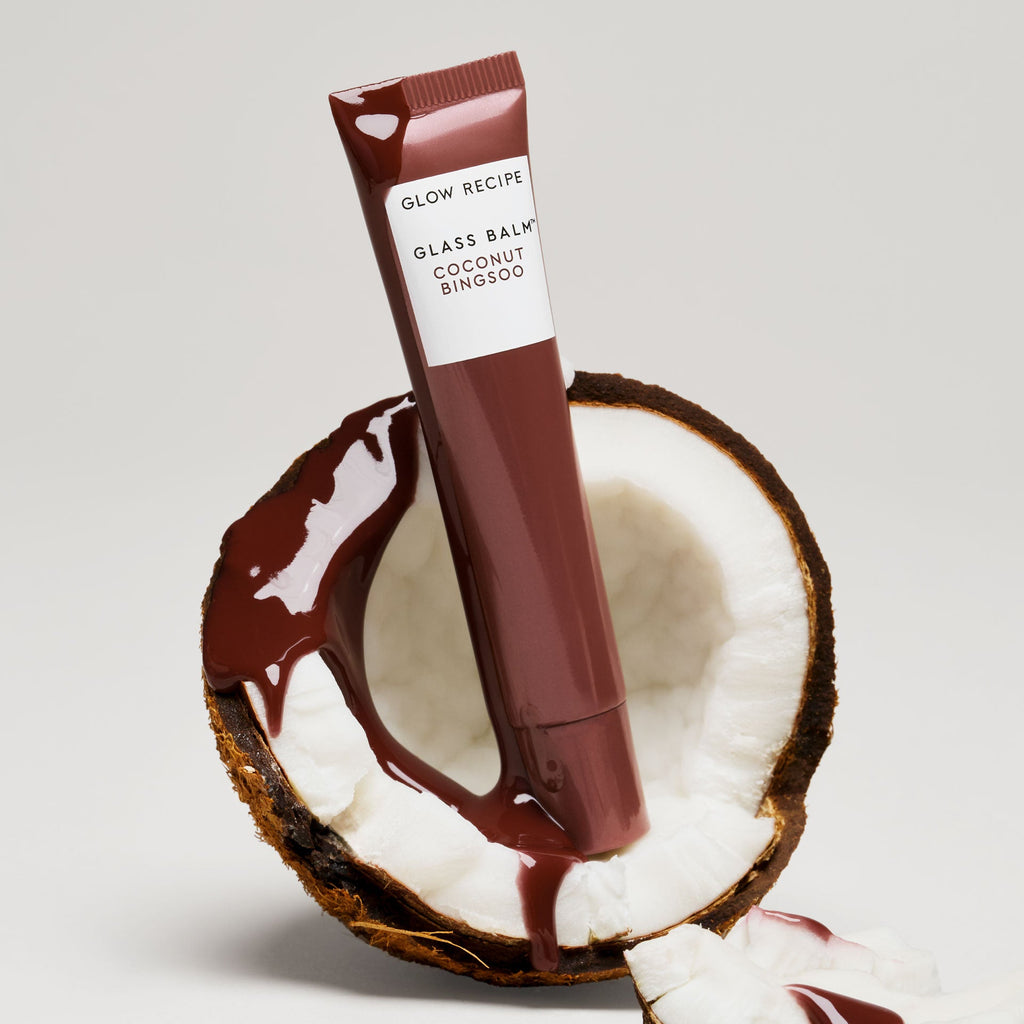 Glass Balm tube in Coconut Bingsoo staged beside coconut halves to illustrate the fruit-powered formula.