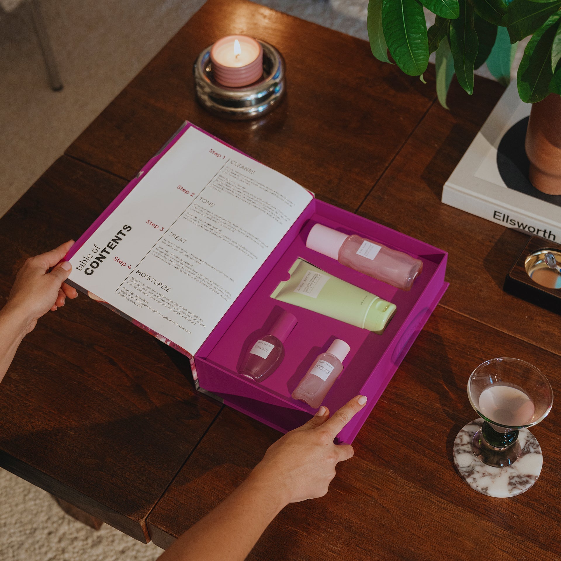 Person opening a pink skin care kit on a wooden table with a candle and plant in the background.
