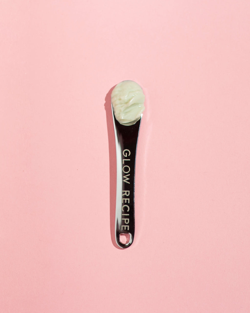 Glow Recipe silver skin care spatula with cream product in scoop on pink background.
