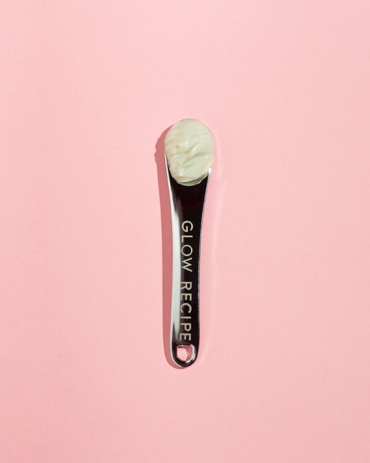 Glow Recipe silver skin care spatula with cream product in scoop on pink background.