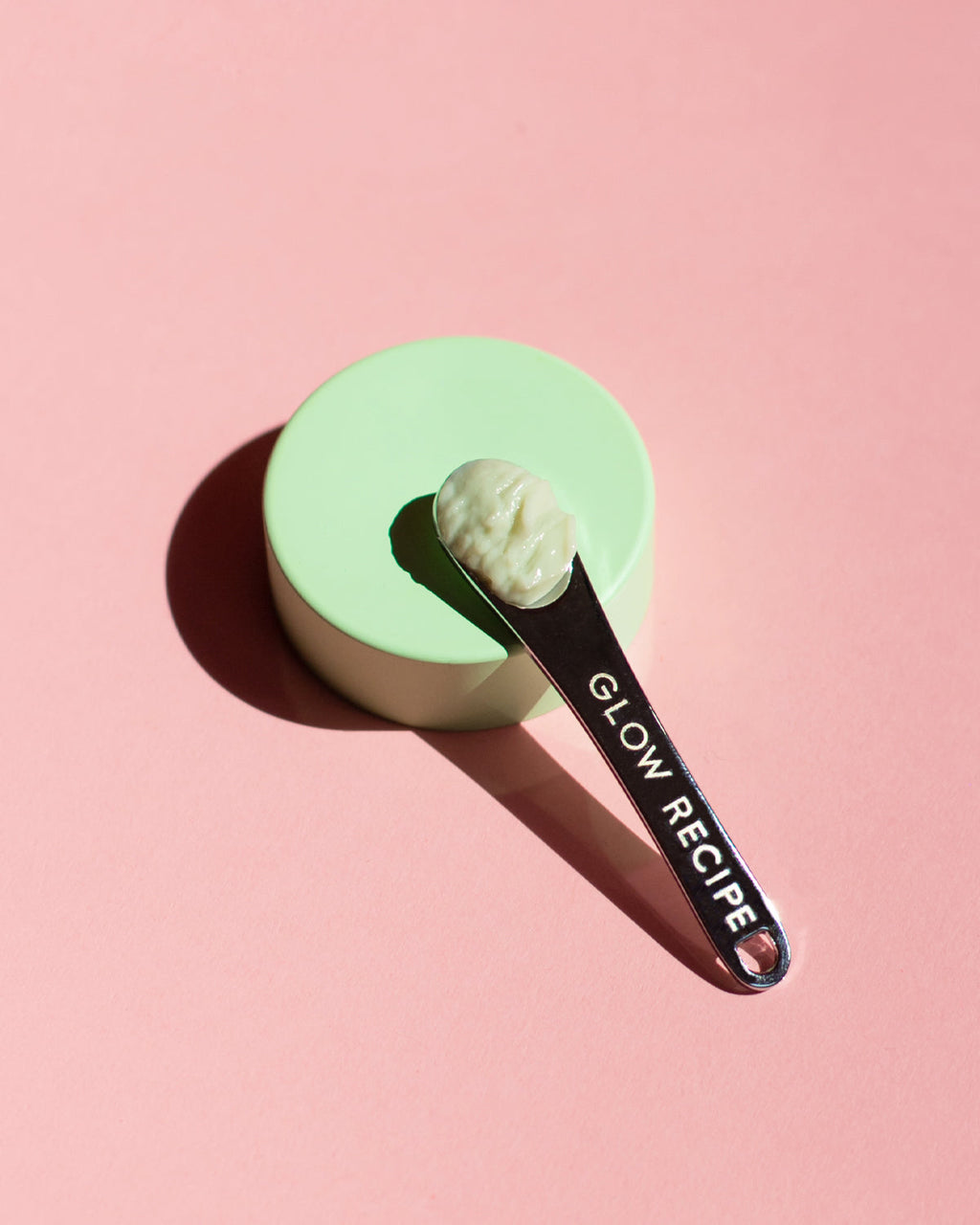 Glow Recipe silver skin care spatula with cream in bowl n top of green stand with pink background.