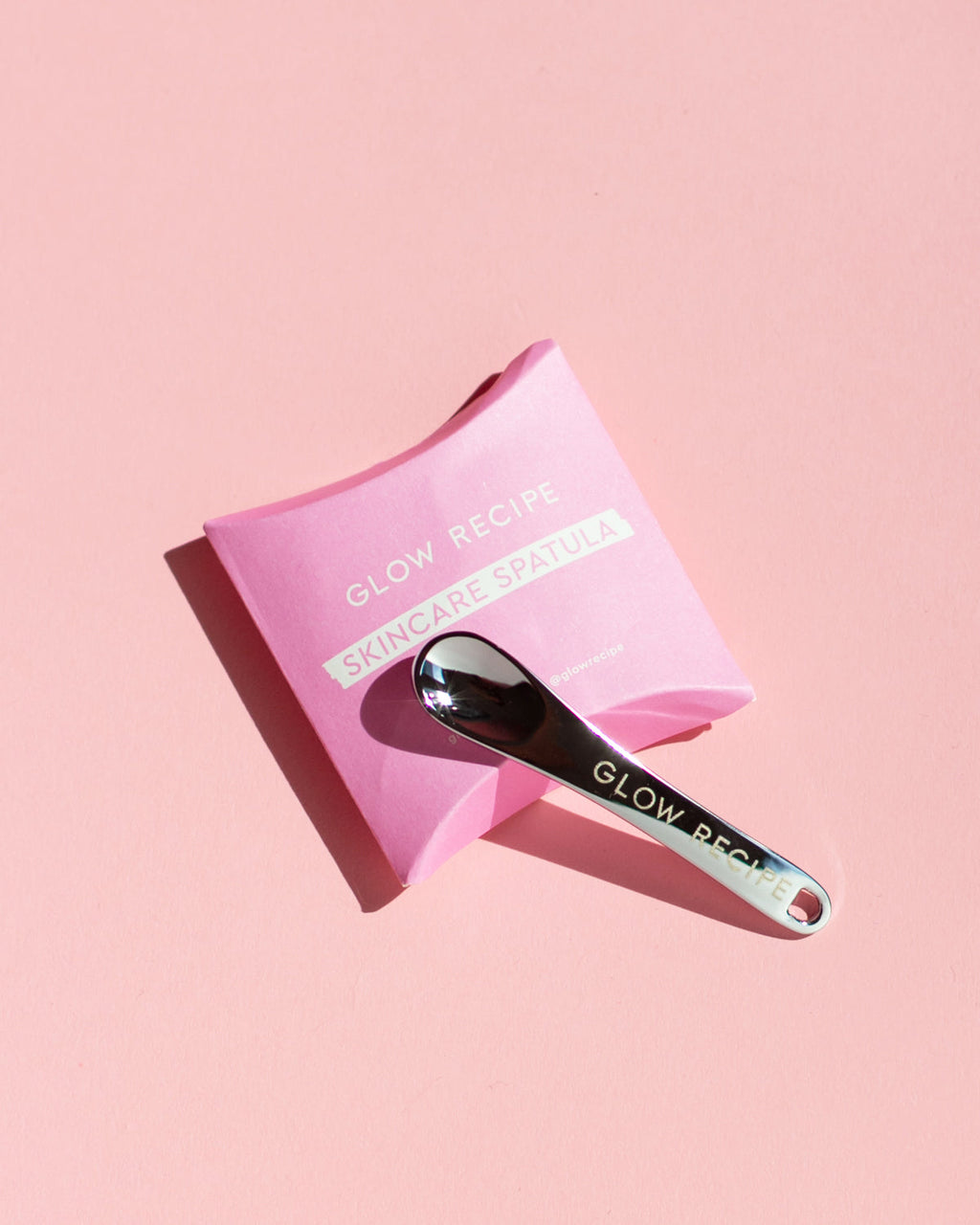 Pink pouch with a skin care spatula on a pink background, featuring 'Glow Recipe' branding.