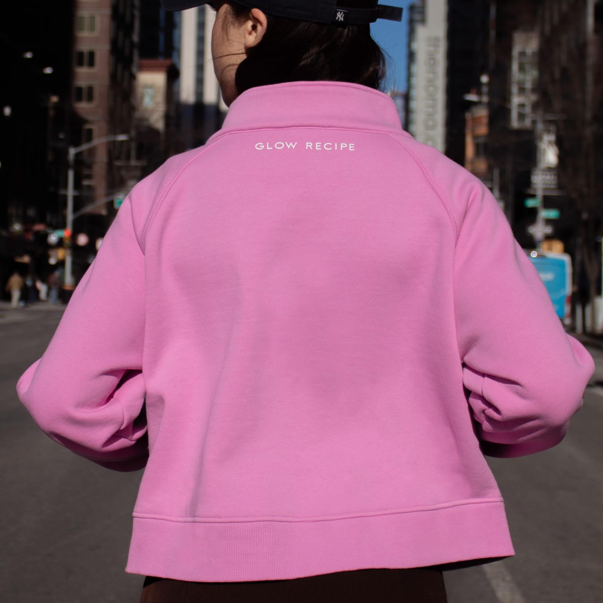 Woman wearing pink zip up Glow recipe written in white script at the neck line. She is walking down a city street.