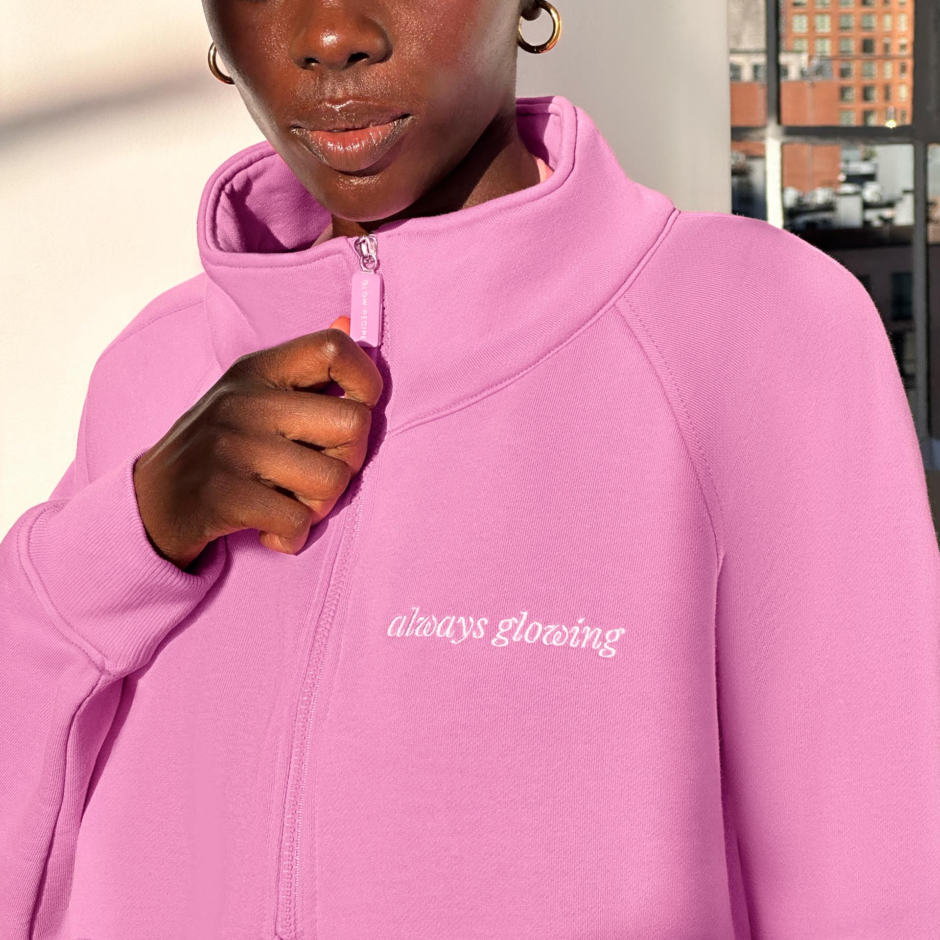 Woman wearing a pink zip up with "always glowing" written in white script on the left breast. She is holding the zipper.