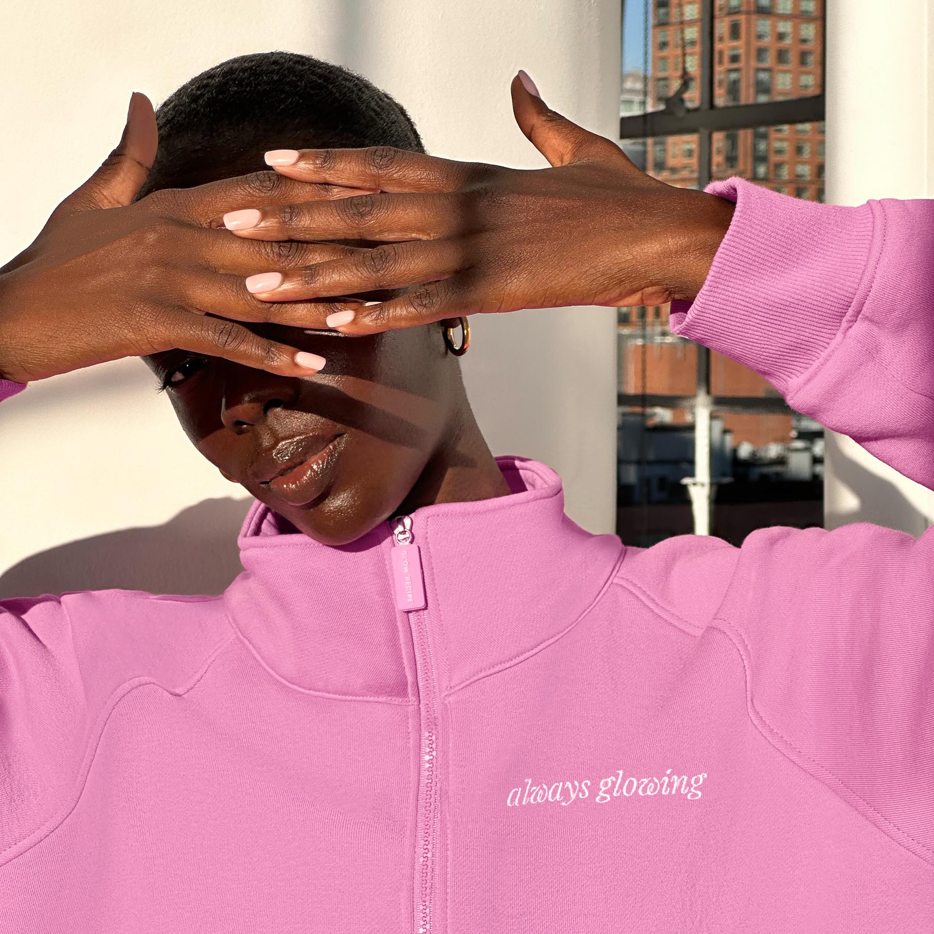 A woman wearing a pink zip up with "Always Glowing" written in white cursive script on the left breast.